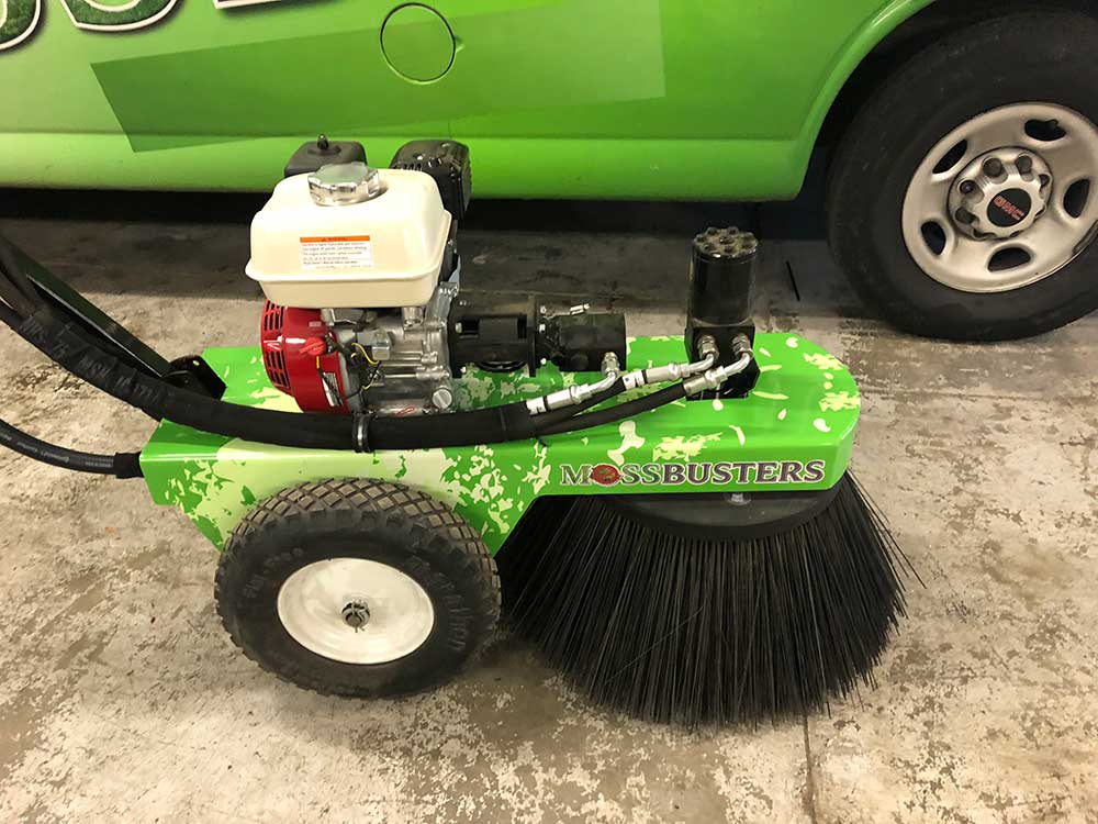 A green mossbusters-branded mechanical brush for moss removal, parked next to a vehicle in a garage.