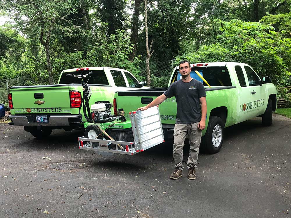 A man standing next to a green truck labeled "job busters" with lawn care equipment loaded at the back, in a wooded area.