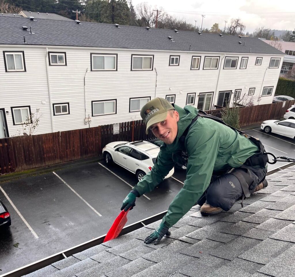 A man in a green jacket and baseball cap uses a red tool to work on a shingled rooftop, with parked cars and a building in the background.