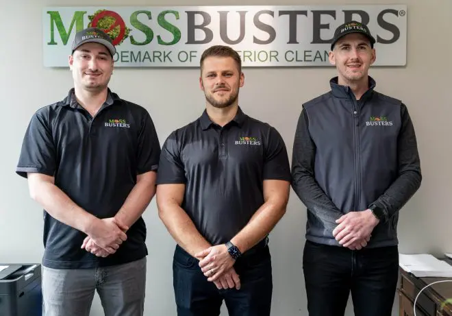 Three men in uniform with "Professional Roof Cleaning Portland OR" logos on their caps and shirts, standing side by side and smiling in an office setting.
