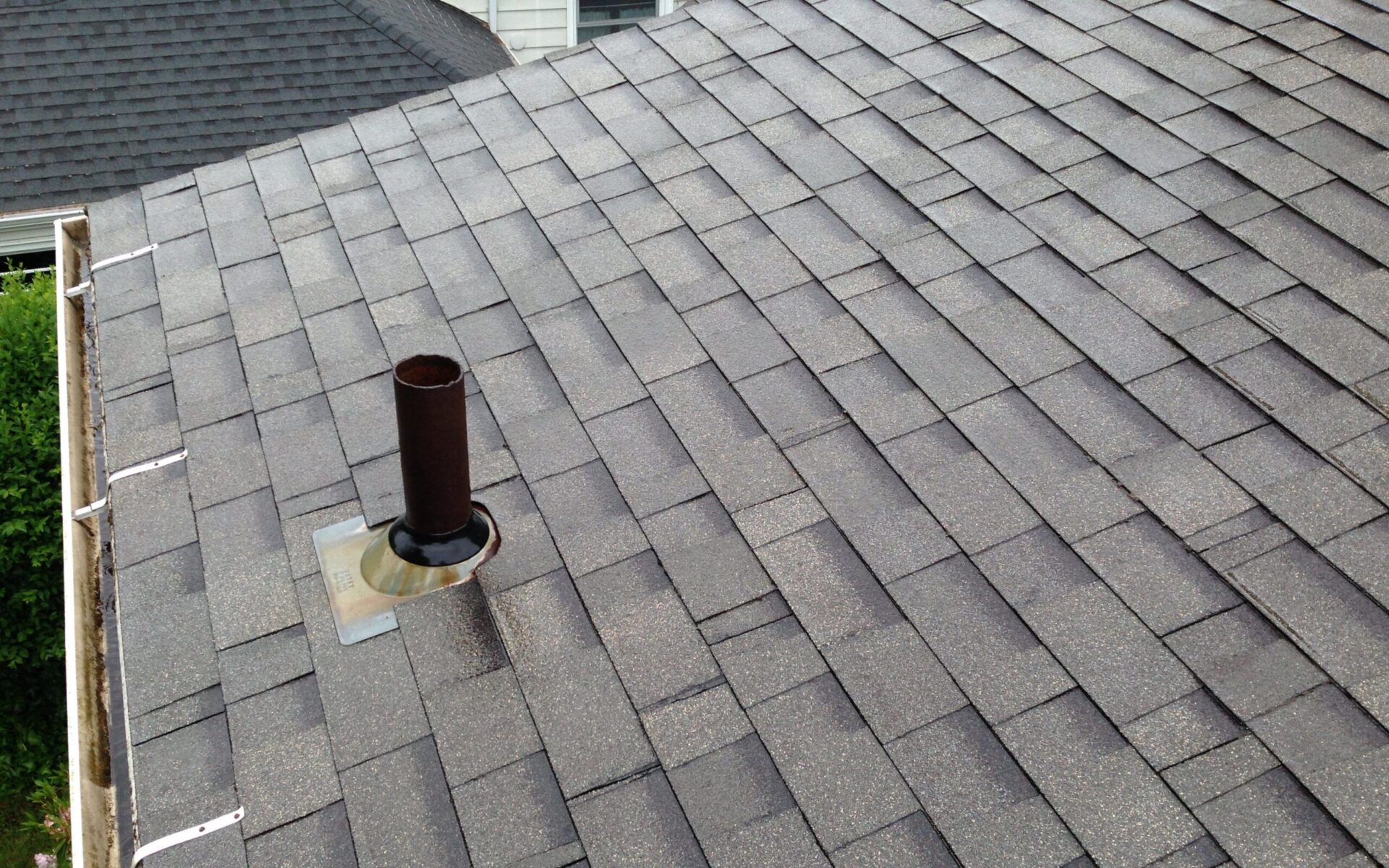 A close-up view of a gray shingled roof with a metal vent pipe, surrounded by a square base, seen under overcast skies.