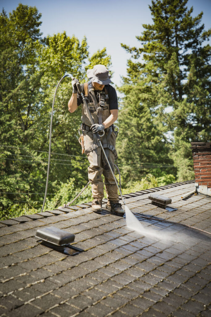A worker applying moss treatment in Portland, OR, using a high-pressure hose to clean a shingled rooftop, wearing safety harness and helmet, surrounded by trees.